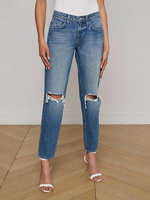 A person wearing L'AGENCE Nevia Slouchy Straight-Leg Jeans with knee rips, a white top, and white heeled sandals stands on a wooden floor against a plain light-colored wall.