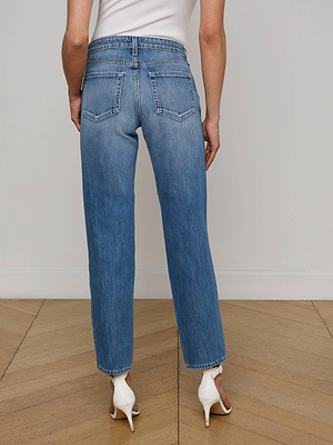 A person stands on a wooden floor, facing away, wearing L'AGENCE Nevia Slouchy Straight-Leg Jeans in vintage-inspired blue wash with white heeled sandals and arms relaxed by their sides.