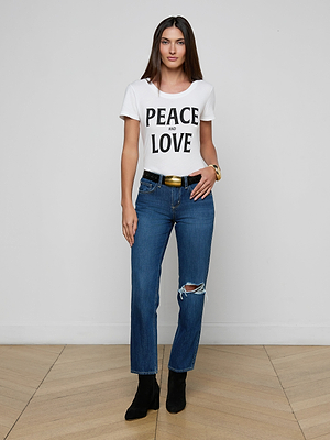 A woman stands on a wooden floor against a white wall, wearing a white PEACE AND LOVE t-shirt, Nevia Slouchy Straight-Leg Jeans with a ripped knee, a black belt, black boots, and a gold bracelet.