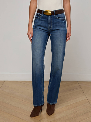 Wearing L'AGENCE Jones Stovepipe Jean in blue, a white sleeveless top, a brown belt with a gold buckle, and brown pointed-toe boots, a person stands on a wooden floor against a plain light-colored wall.