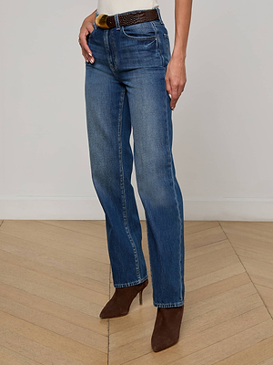 A person wearing the L'AGENCE Jones Stovepipe Jean in all-cotton denim, styled with a brown belt and heeled ankle boots, stands on a wooden floor against a plain light-colored wall.