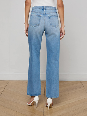 A person wearing L'AGENCE Jones Stovepipe Jeans and white high-heeled shoes stands on a wooden floor, photographed from the back.