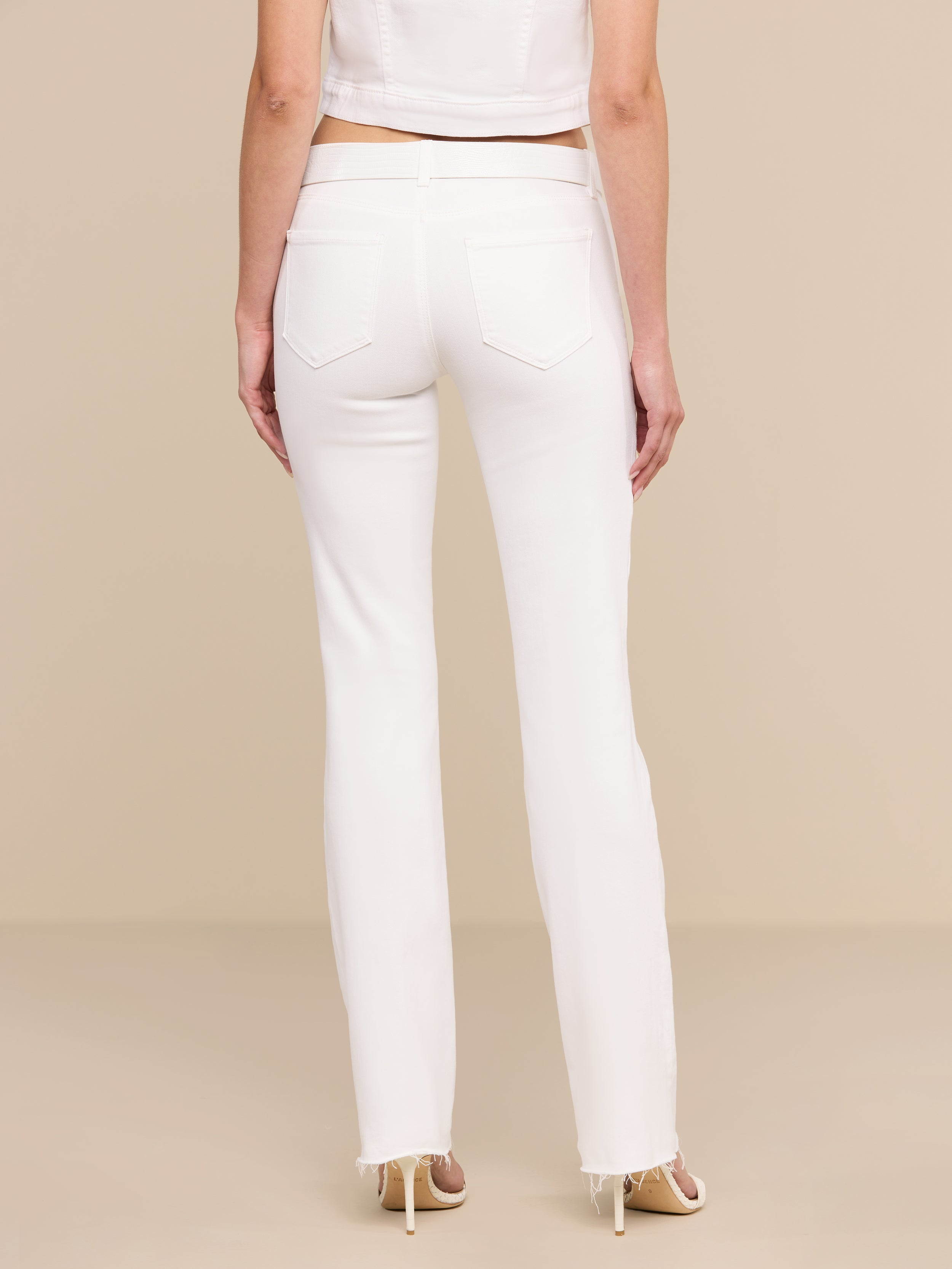 A woman stands facing away, wearing the Sneeki Low-Rise Straight-Leg Jean in white with a raw hem, paired with a white cropped top and beige heels against a plain white background.