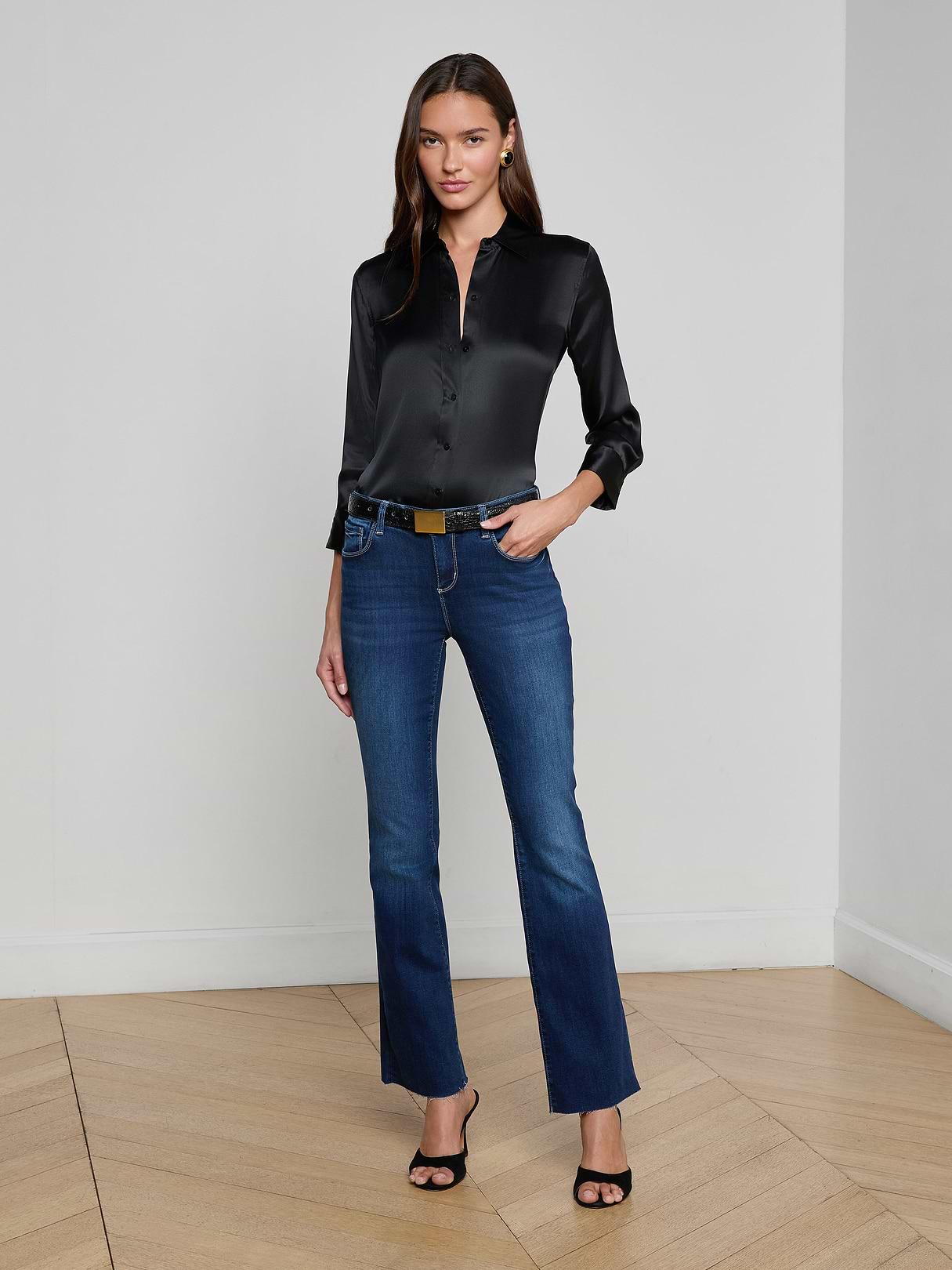 A woman poses indoors against a plain wall, wearing L'AGENCE Sneeki Low-Rise Straight-Leg Jeans with a black satin button-up blouse, black heeled sandals, and a black belt with a gold buckle. She stands on a wooden floor with one hand in her pocket.