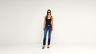 A woman wearing sunglasses, a black tank top, blue jeans, and black heels stands against a plain white background.