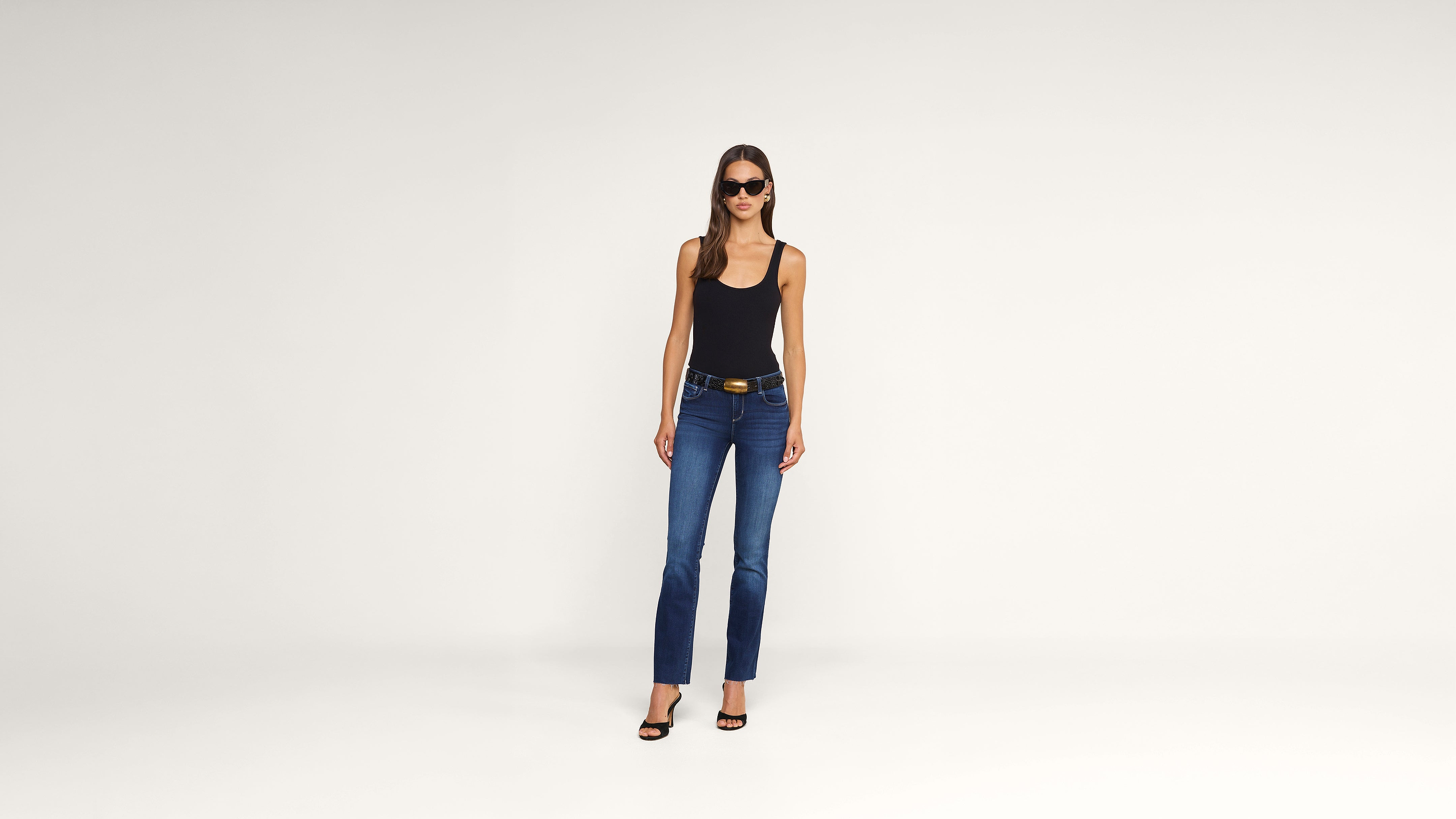 A woman wearing sunglasses, a black tank top, blue jeans, and black heels stands against a plain white background.