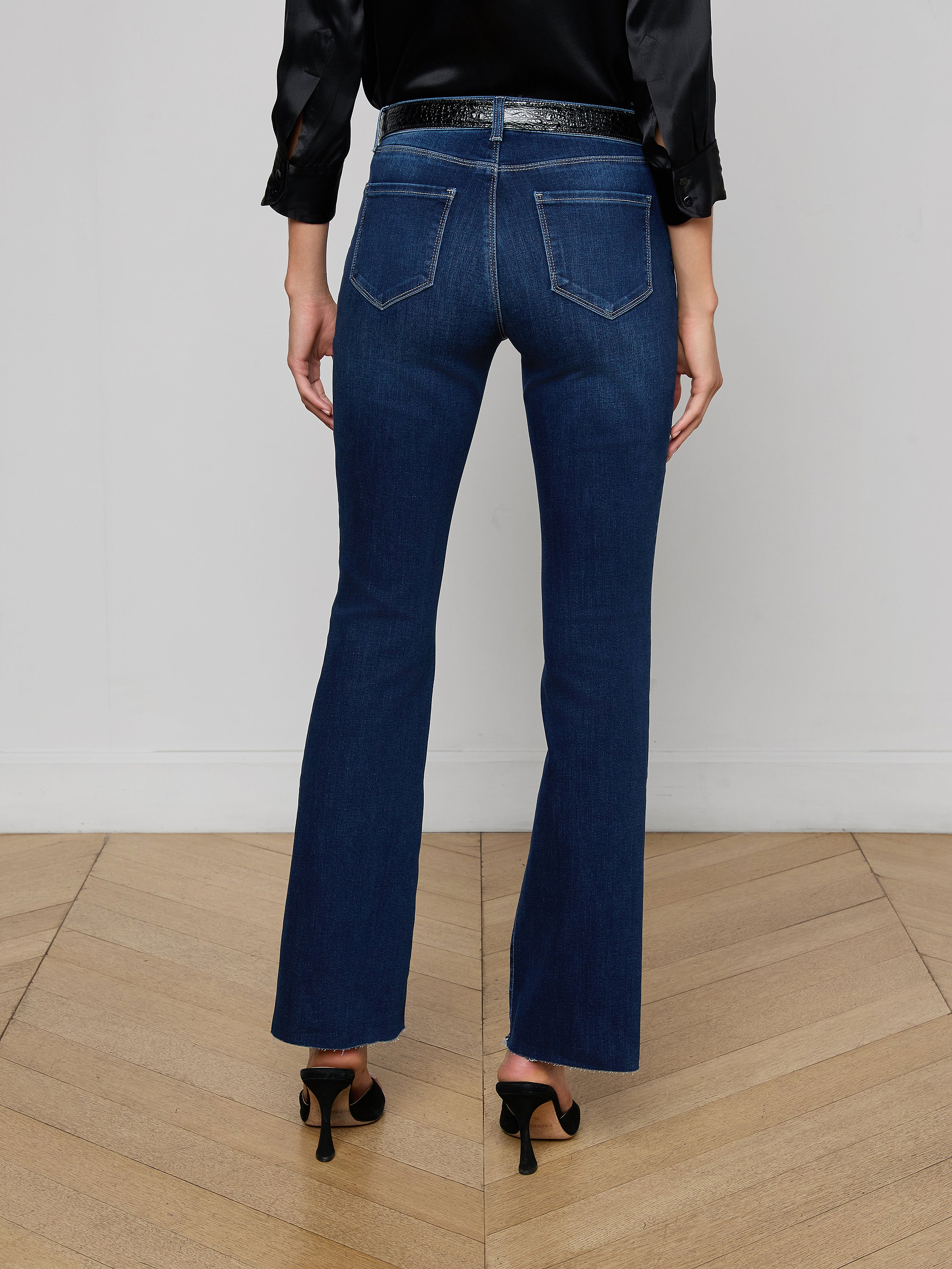 A person stands facing away, wearing a black sleeveless top, a pair of Sneeki Low-Rise Straight-Leg Jeans in dark wash super stretch denim with back pockets, a black belt, and black heeled shoes against a plain white background.