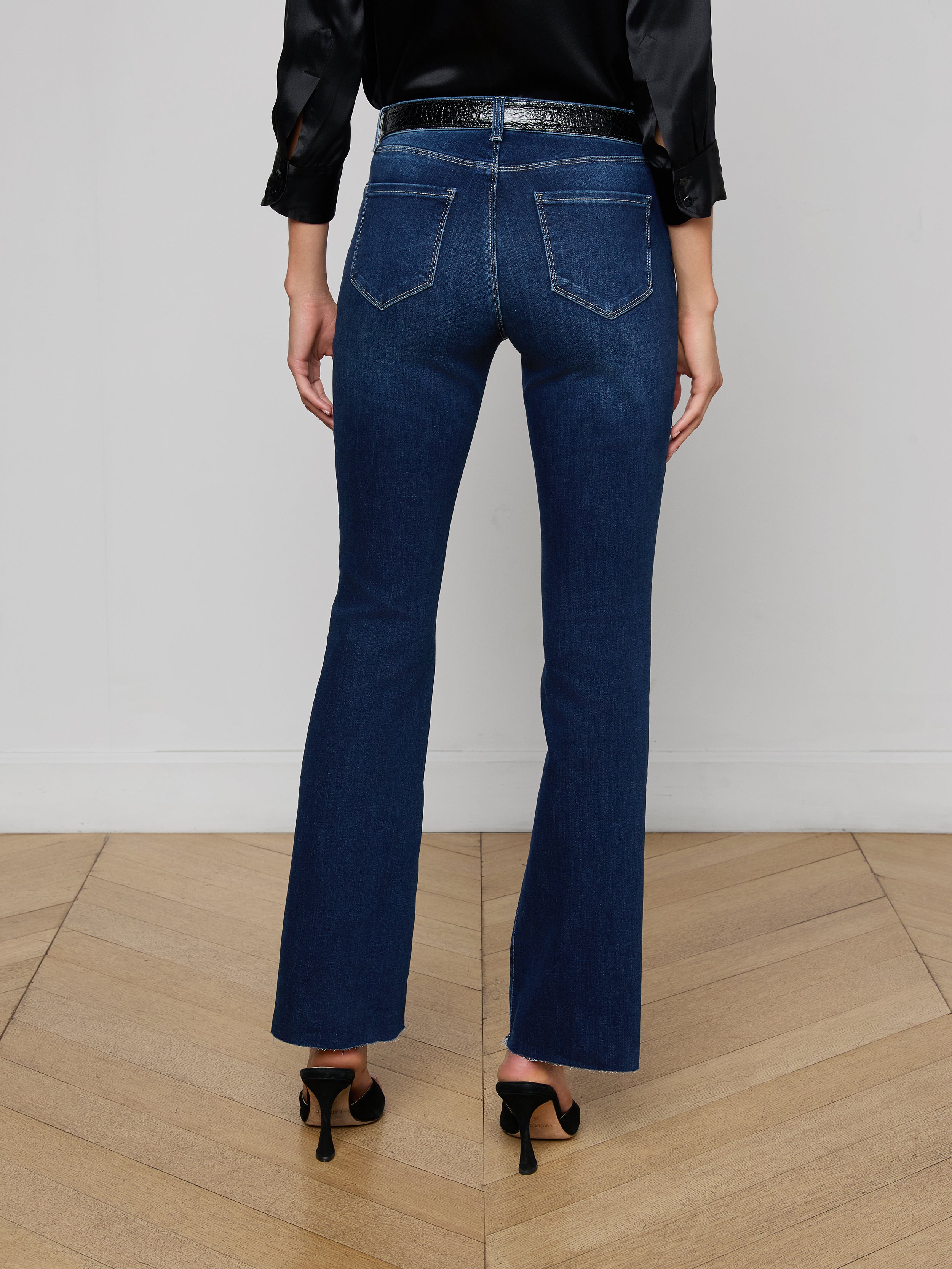 A person stands facing away, wearing a black sleeveless top, a pair of Sneeki Low-Rise Straight-Leg Jeans in dark wash super stretch denim with back pockets, a black belt, and black heeled shoes against a plain white background.