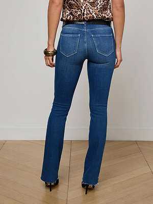 A person stands with their back to the camera on a wooden floor, wearing L'AGENCE Sneeki Low-Rise Straight-Leg Jeans in blue, black high heels, a leopard print top, and a chunky bracelet.