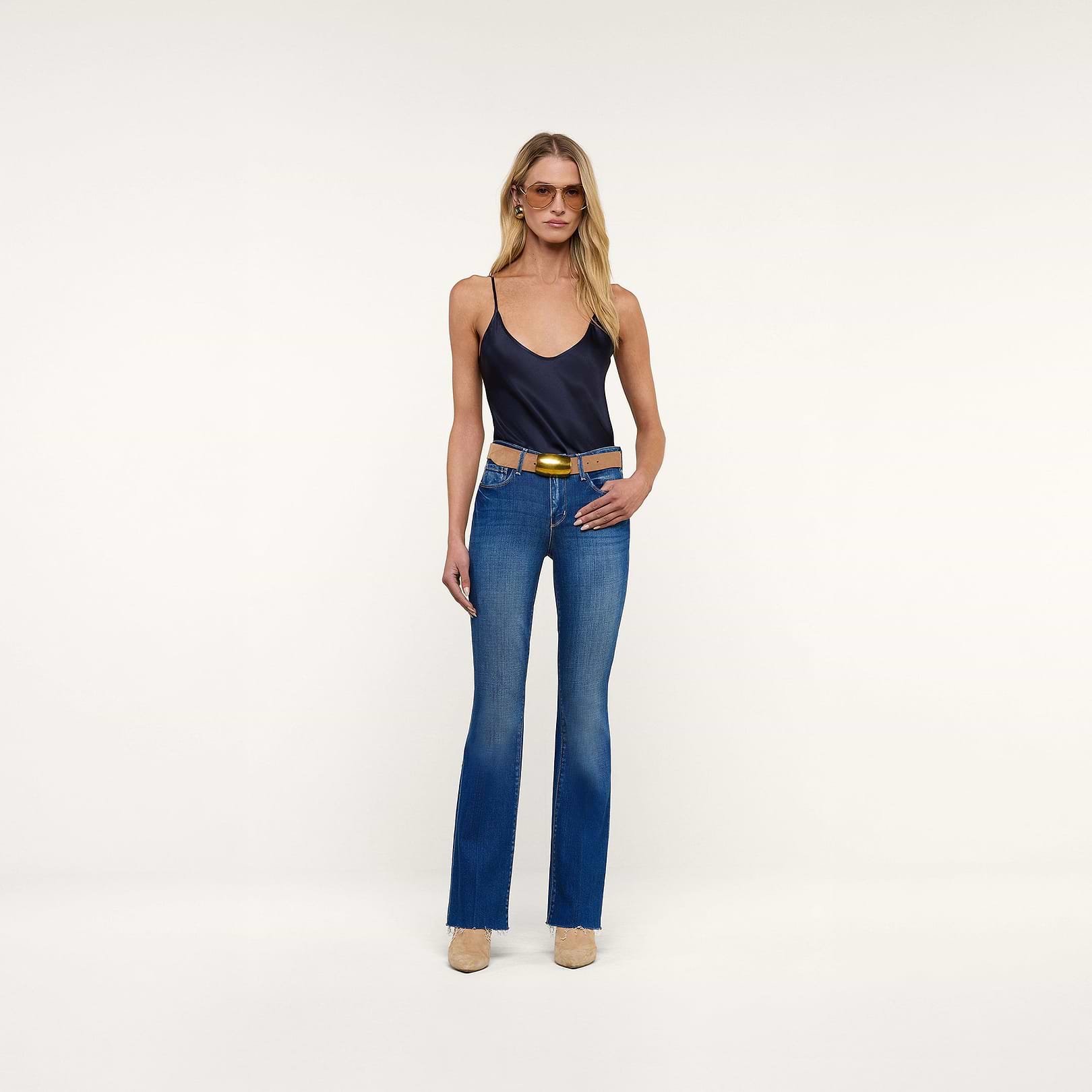 A woman stands against a plain white background wearing a navy camisole, the Sneeki Low-Rise Straight-Leg Jean in blue, a tan belt, sunglasses, and nude heels. She faces forward with one hand on her hip.