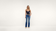 A woman stands facing forward, wearing a black sleeveless top, blue jeans, beige boots, and a brown belt, set against a plain, light background.