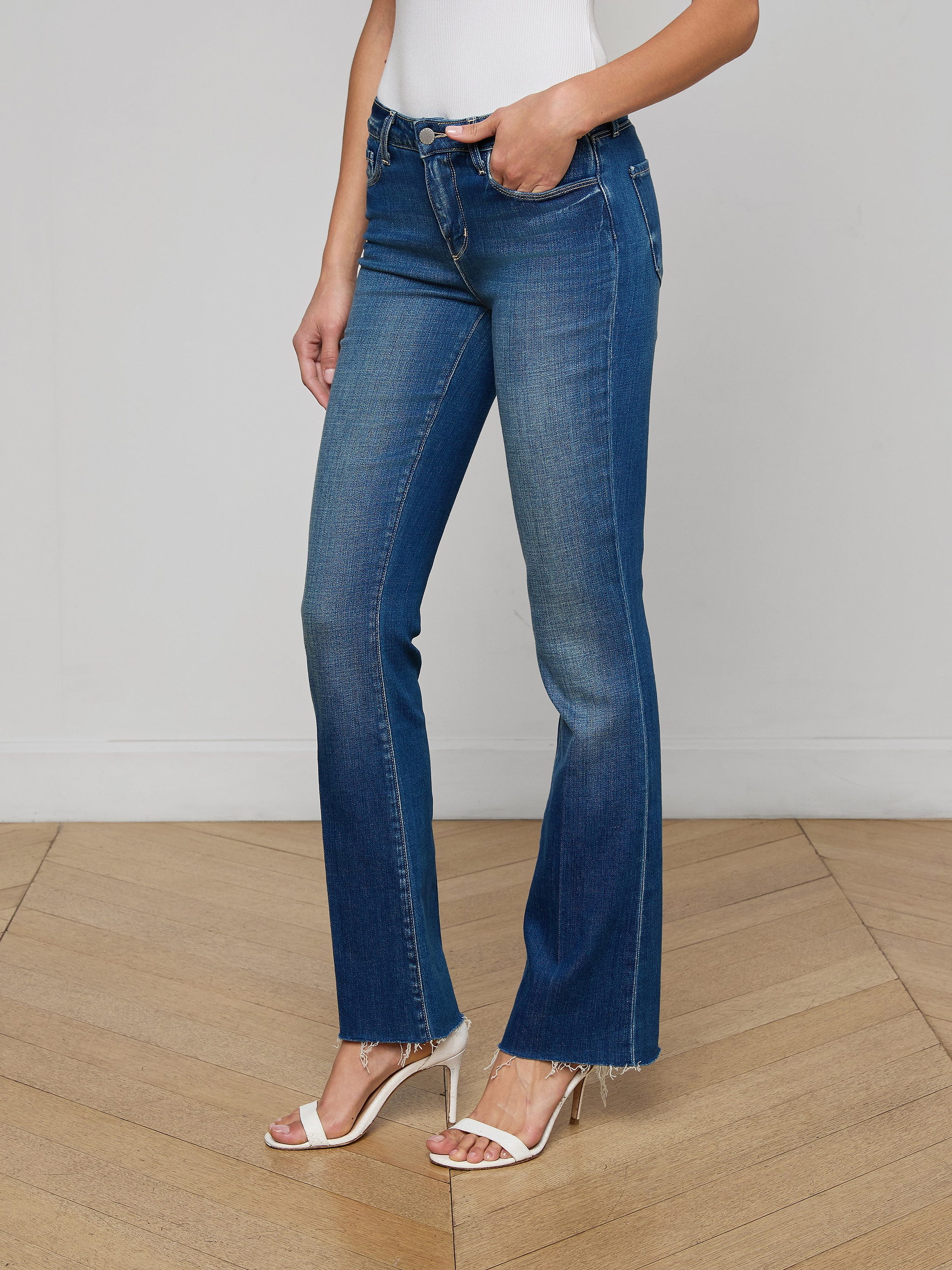 A woman stands against a plain white background wearing Sneeki Low-Rise Straight-Leg Jeans in blue with frayed hems, styled with a brown belt, a dark sleeveless top, and beige heeled shoes.