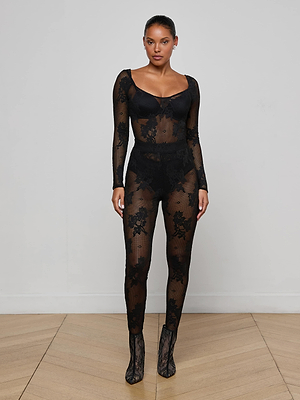 A woman stands indoors against a plain wall, wearing the Josiane Floral Lace Legging with a sheer black lace bodysuit and lace high-heeled shoes. She faces forward with a neutral expression.