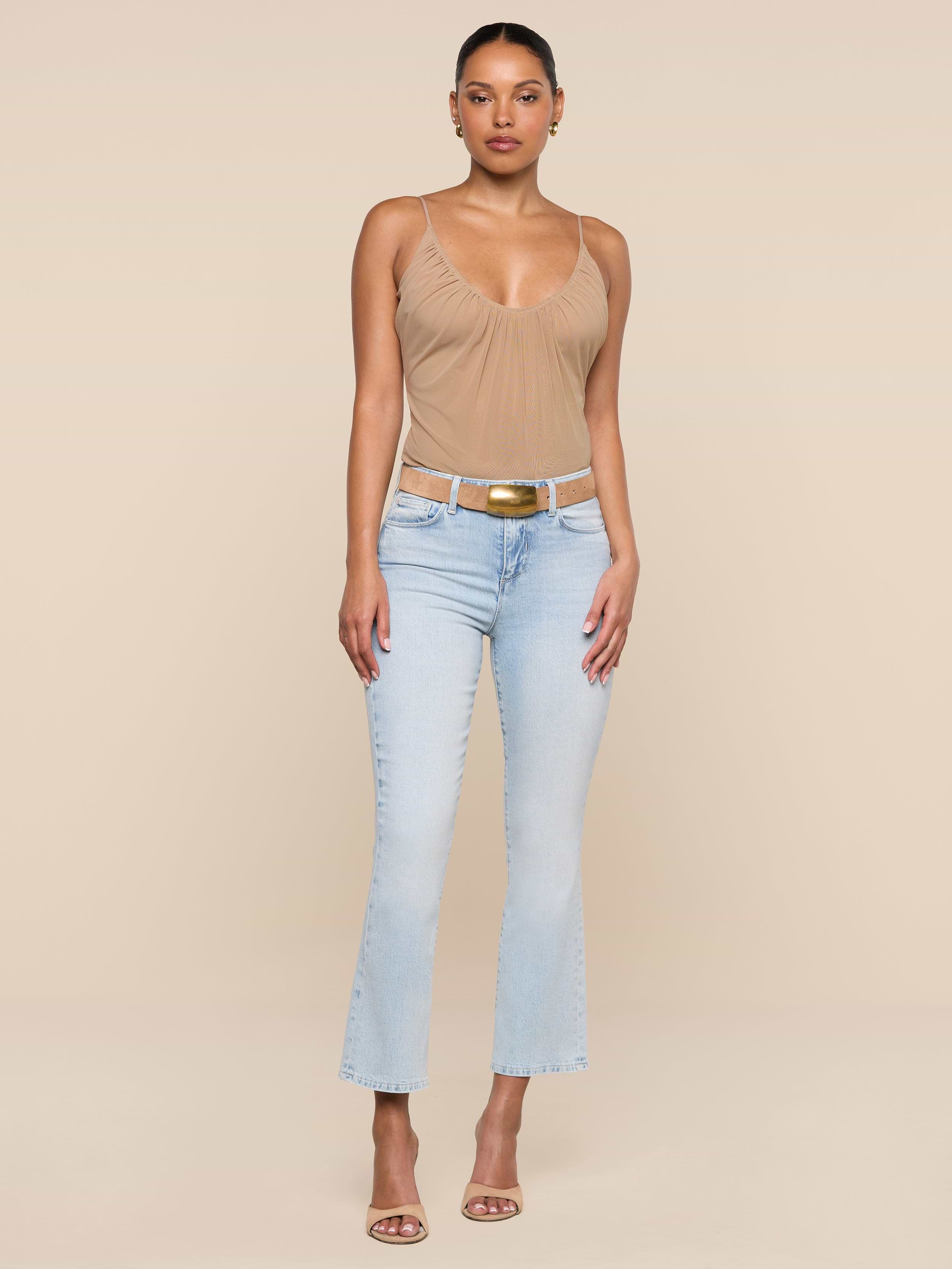 A woman faces forward against a beige background wearing the Tati Cropped Micro Boot Jean light wash, a tan sleeveless top, brown belt with gold buckle, and beige heeled sandals. Her hair is pulled back and she has a neutral expression.