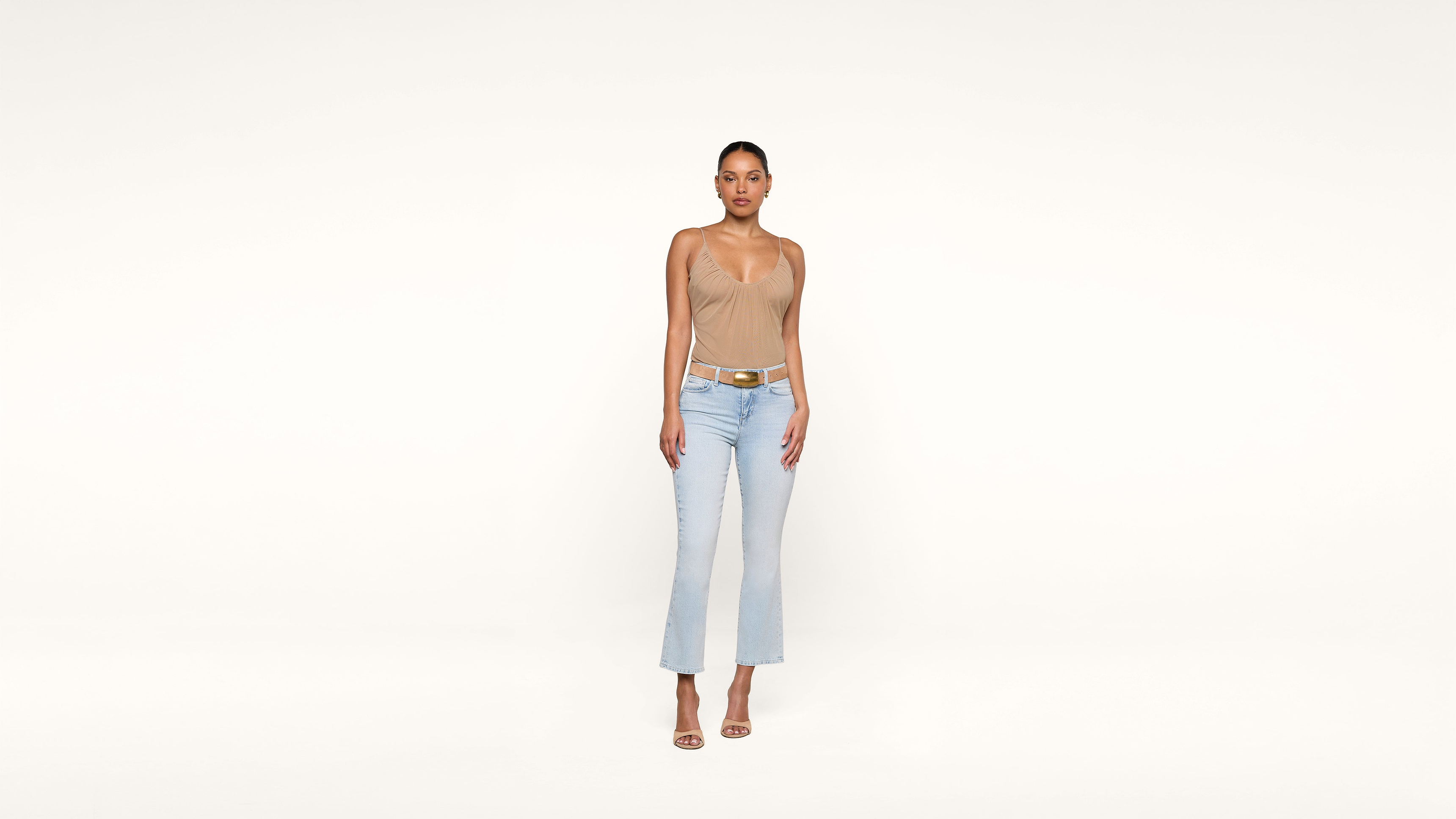 A woman stands against a plain white background, wearing a beige sleeveless top, light blue jeans, and beige high-heeled sandals. She is facing forward with her arms relaxed by her sides.