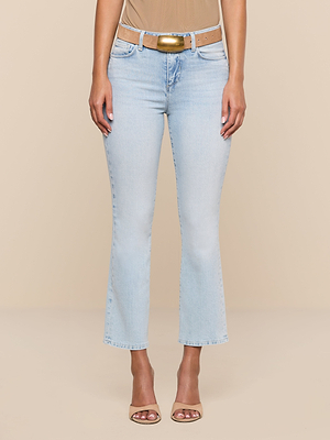 Wearing the Tati Cropped Micro Boot Jean in light blue, paired with a beige belt, nude top, and nude heeled sandals, a person stands against a plain beige background. The image is cropped from shoulders to feet.