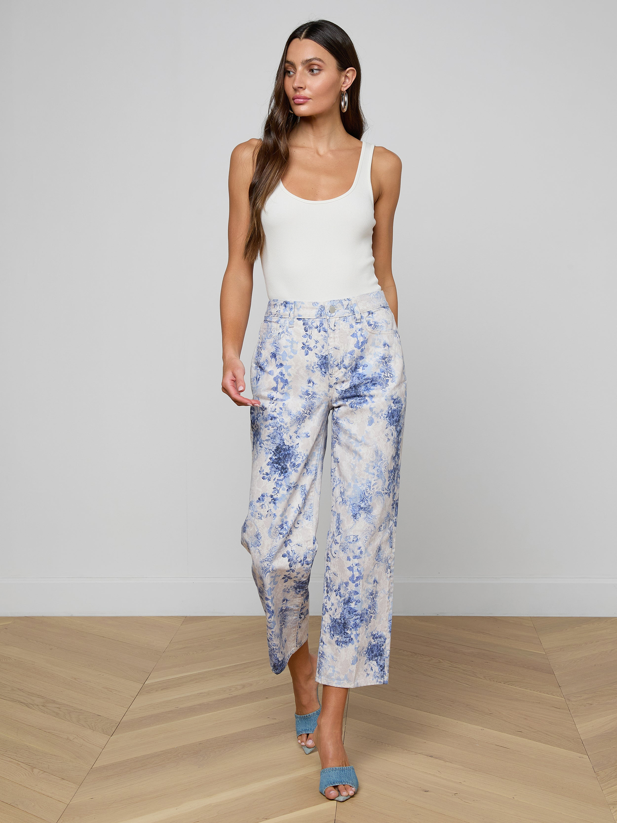 Woman with long brown hair wears a white sleeveless top, June Cropped Stovepipe Jean in a blue and white pattern, and blue heeled sandals while standing against a plain light wall and wooden floor.