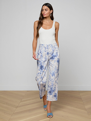 Woman with long brown hair wears a white sleeveless top, June Cropped Stovepipe Jean in a blue and white pattern, and blue heeled sandals while standing against a plain light wall and wooden floor.