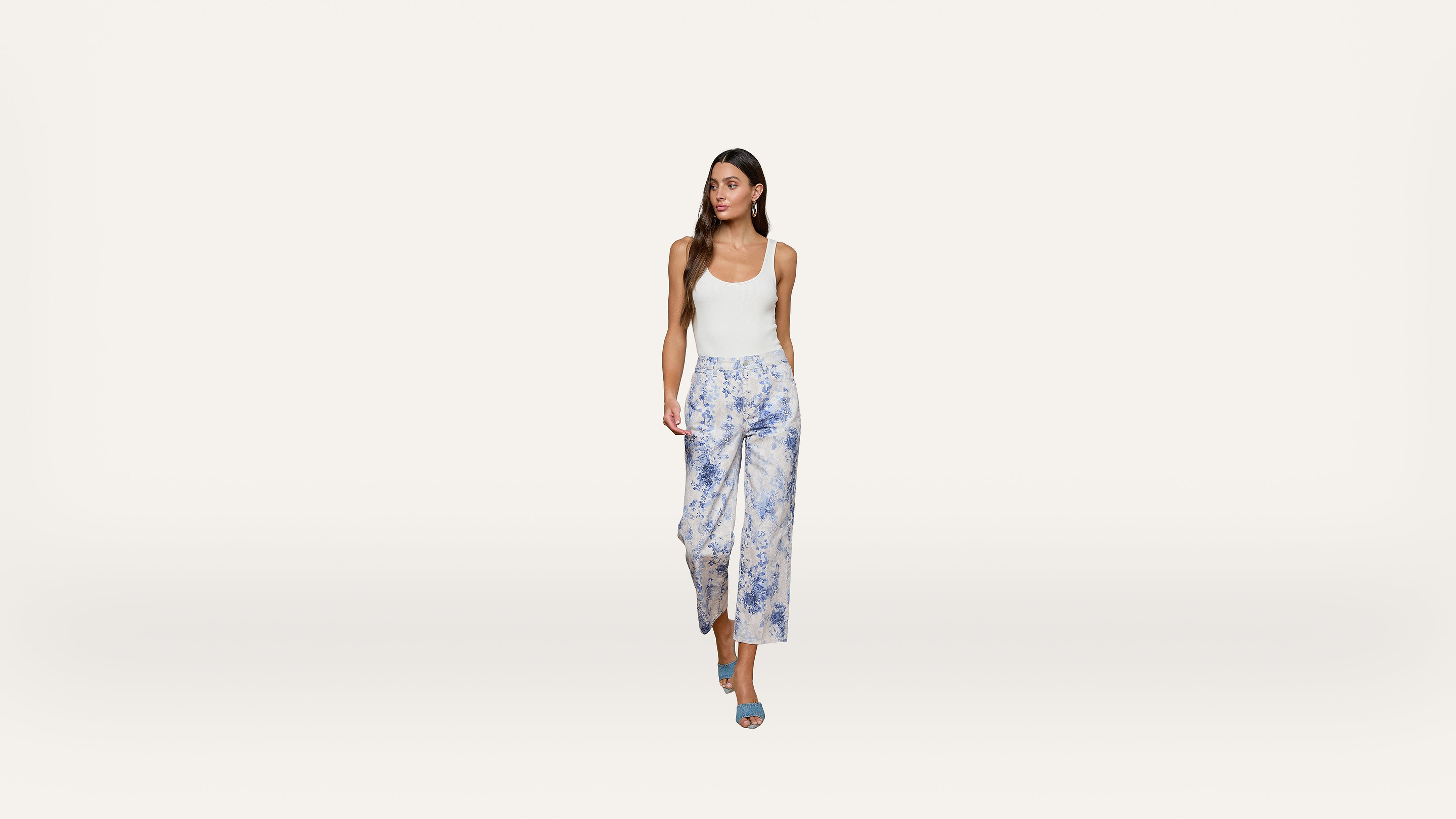Woman in a white tank top, blue patterned wide-leg pants, and blue heels walking forward against a plain white background.