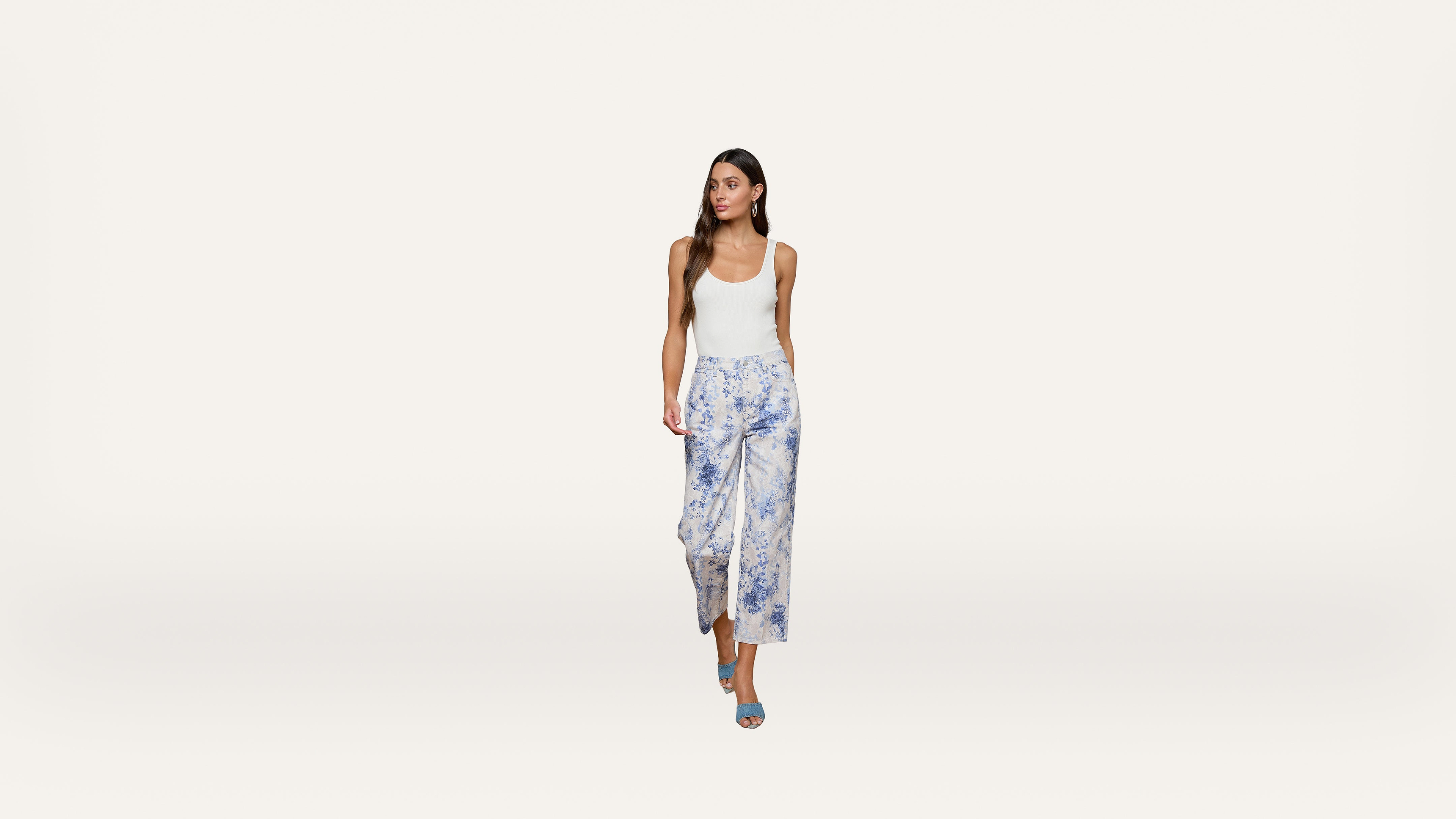 Woman in a white tank top, blue patterned wide-leg pants, and blue heels walking forward against a plain white background.
