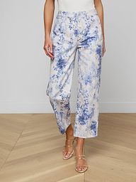 A person wearing June Cropped Stovepipe Jeans in blue and white floral print, a sleeveless white top, and strappy heeled sandals stands on a light wood floor.