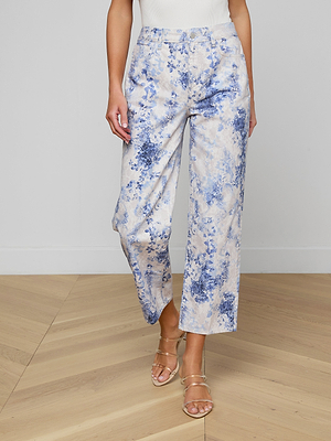 A person wearing June Cropped Stovepipe Jeans in blue and white floral print, a sleeveless white top, and strappy heeled sandals stands on a light wood floor.