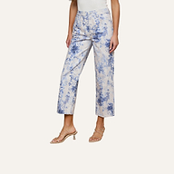 A person wearing June Cropped Stovepipe Jean with a blue and white floral print, paired with a white top and clear heeled sandals, stands on a light wooden floor against a plain white wall.