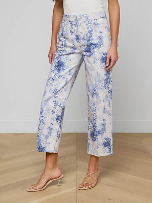 A person wearing June Cropped Stovepipe Jean with a blue and white floral print, paired with a white top and clear heeled sandals, stands on a light wooden floor against a plain white wall.