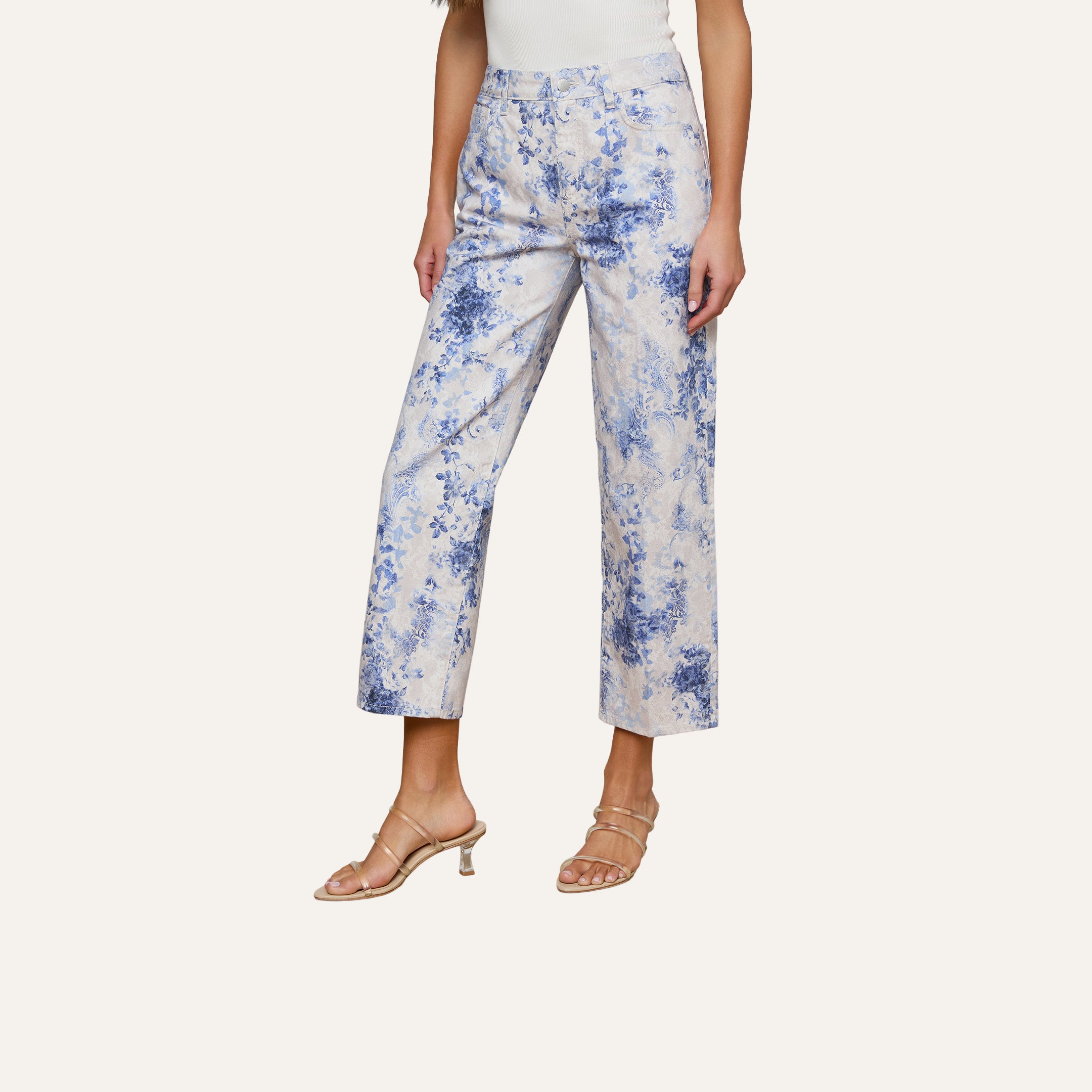 A person wearing June Cropped Stovepipe Jean with a blue and white floral print, paired with a white top and clear heeled sandals, stands on a light wooden floor against a plain white wall.