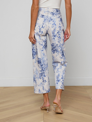 A person stands on a wooden floor, facing away from the camera, wearing June Cropped Stovepipe Jeans with a floral print and clear-heeled sandals.