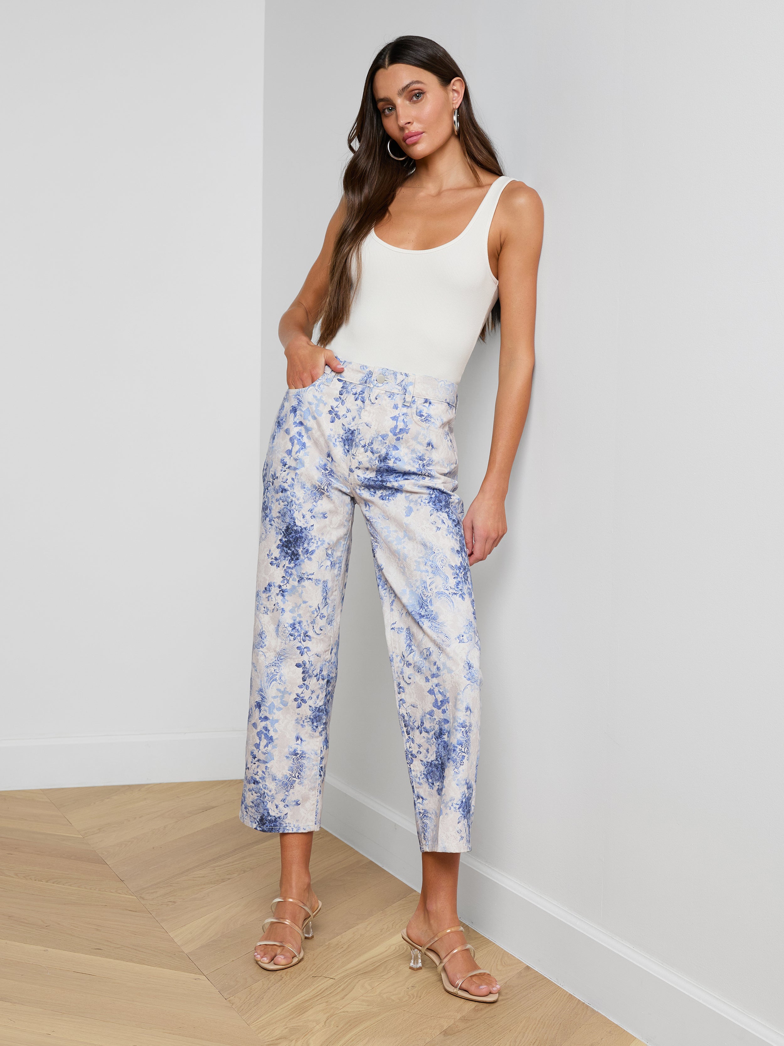 A woman stands indoors against a white wall, wearing a sleeveless white top, strappy heeled sandals, and the June Cropped Stovepipe Jean. She has long brown hair and poses with one hand in her pocket.