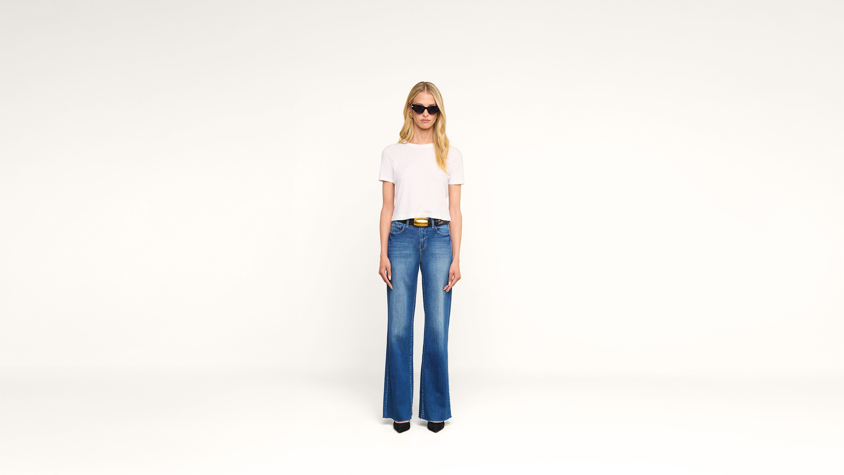 A woman wearing sunglasses, a white t-shirt, blue wide-leg jeans, and black shoes stands against a plain white background.