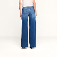 A person wearing a white top and the Scottie Wide-Leg Jean in vintage-wash blue denim, featuring high-rise, wide-leg silhouette with frayed hems and visible back pockets, stands with their back to the camera against a plain white background.