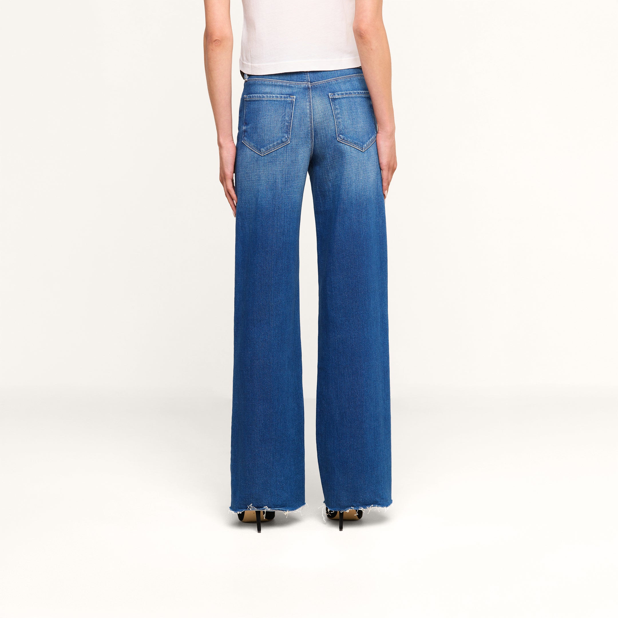 A person wearing a white top and the Scottie Wide-Leg Jean in vintage-wash blue denim, featuring high-rise, wide-leg silhouette with frayed hems and visible back pockets, stands with their back to the camera against a plain white background.