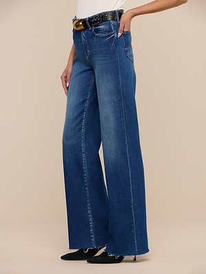 A person wears the Scottie Wide-Leg Jean in a vintage wash with frayed hems, styled with a black belt and black pointed-toe shoes, standing against a beige background. Their upper body is mostly out of frame.