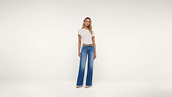 A woman stands against a plain white background, wearing a fitted white short-sleeve top, wide-leg blue jeans, a brown belt, and beige shoes.