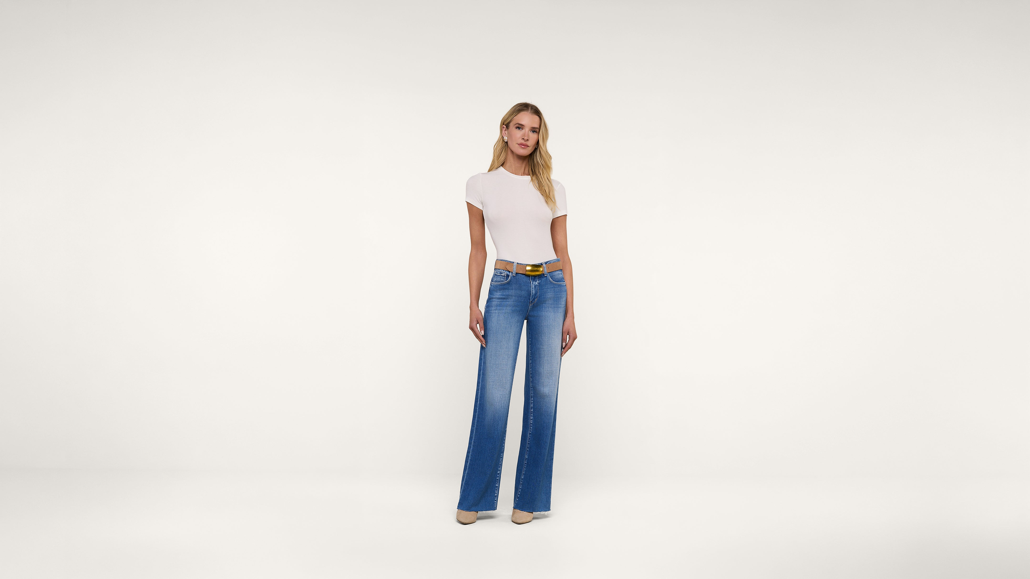 A woman stands against a plain white background, wearing a fitted white short-sleeve top, wide-leg blue jeans, a brown belt, and beige shoes.