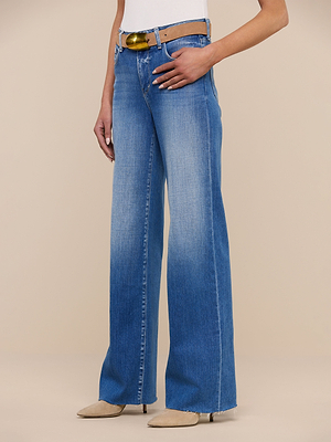 A person wearing Scottie Wide-Leg Jeans in a blue faded wash with a raw hem, a yellow-brown belt, white top, and beige pointed-toe shoes stands against a plain beige background.