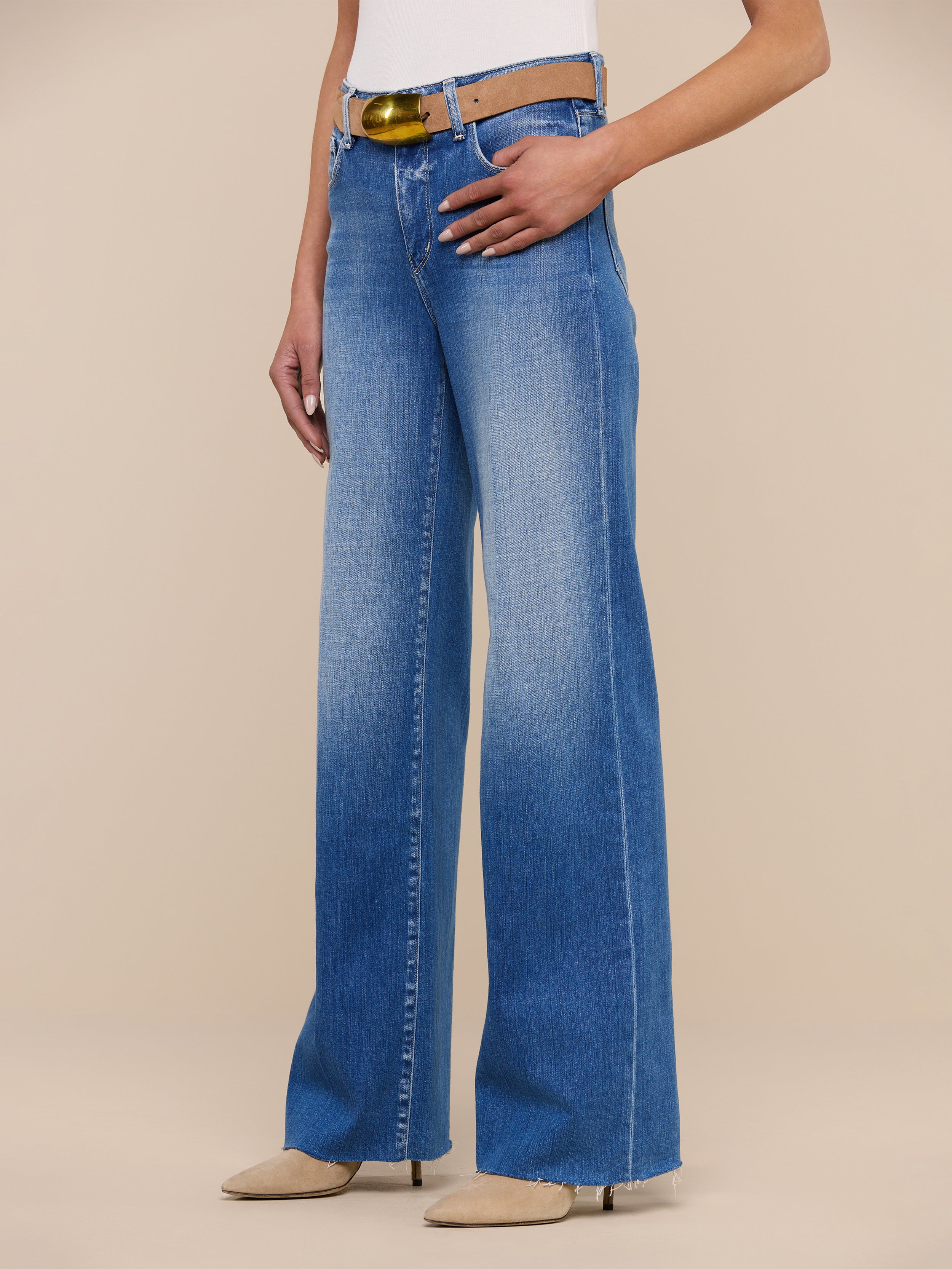 Wearing the Scottie Wide-Leg Jean with a high waist and raw hem, styled with a light brown belt and beige pointed-toe shoes, standing against a plain white background. Upper body is partially out of frame.