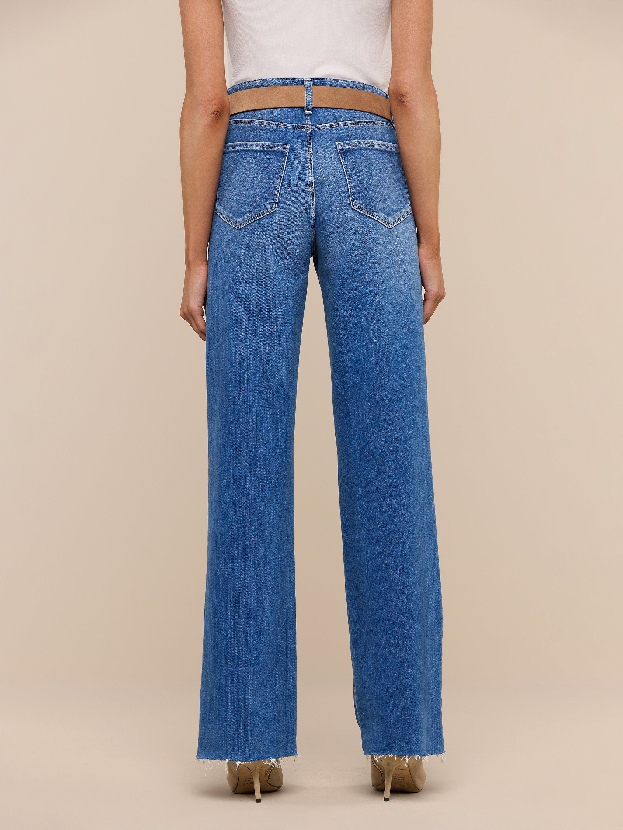 A person wearing a white top and the Scottie Wide-Leg Jean in a blue high-rise style stands facing away, showcasing the back pocket and seam details. The background is plain and light-colored.