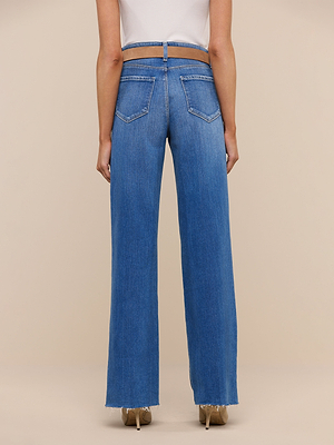 Wearing Scottie Wide-Leg Jean—high-rise with a unique open-back waistband, visible pockets, and raw hems—a person pairs them with a white top, standing in front of a plain beige background.