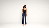 A woman wearing sunglasses, a black tank top, wide-leg blue jeans, and black heels stands against a plain white background.