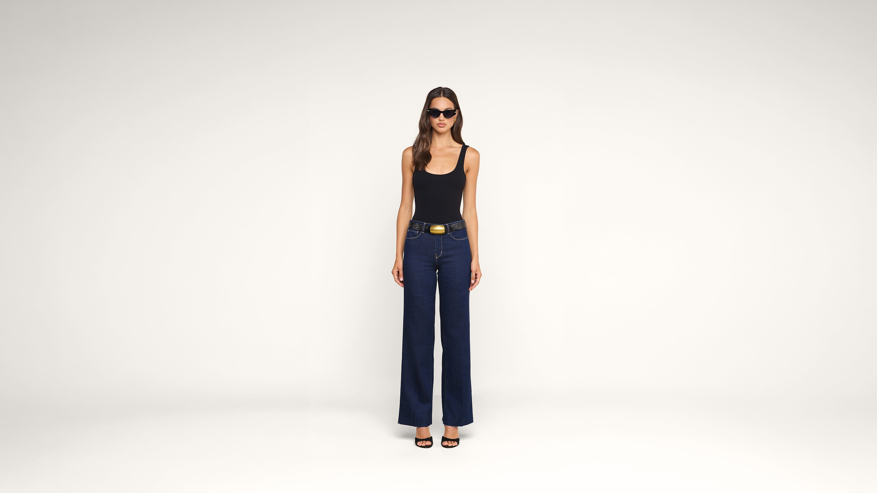 A woman wearing sunglasses, a black tank top, wide-leg blue jeans, and black heels stands against a plain white background.