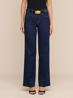 Une personne portant un jean Scottie à jambes larges, une ceinture noire ornée dune grande boucle dorée, un haut noir et des escarpins noirs à bout ouvert se tient debout devant un fond beige.