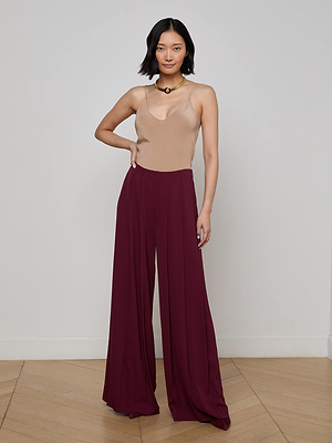 A person stands indoors on a wooden floor, wearing a beige sleeveless top and the L'AGENCE Nova Pleated Wide-Leg Pant in maroon. They have medium-length dark hair and pose with one hand on their hip against a plain light wall.