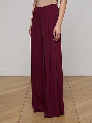 A person wears the L'AGENCE Nova Pleated Wide-Leg Pant in maroon and a beige top, standing on a light wood floor against a plain light wall. Only the lower half of their body is visible.