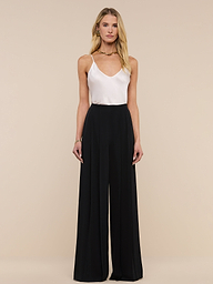 A woman stands against a plain white background, wearing a white sleeveless top paired with the Nova Pleated Wide-Leg Pant, a high-waisted tailored black trouser. Her outfit features wide-leg trousers. She has long blonde hair and a neutral expression.