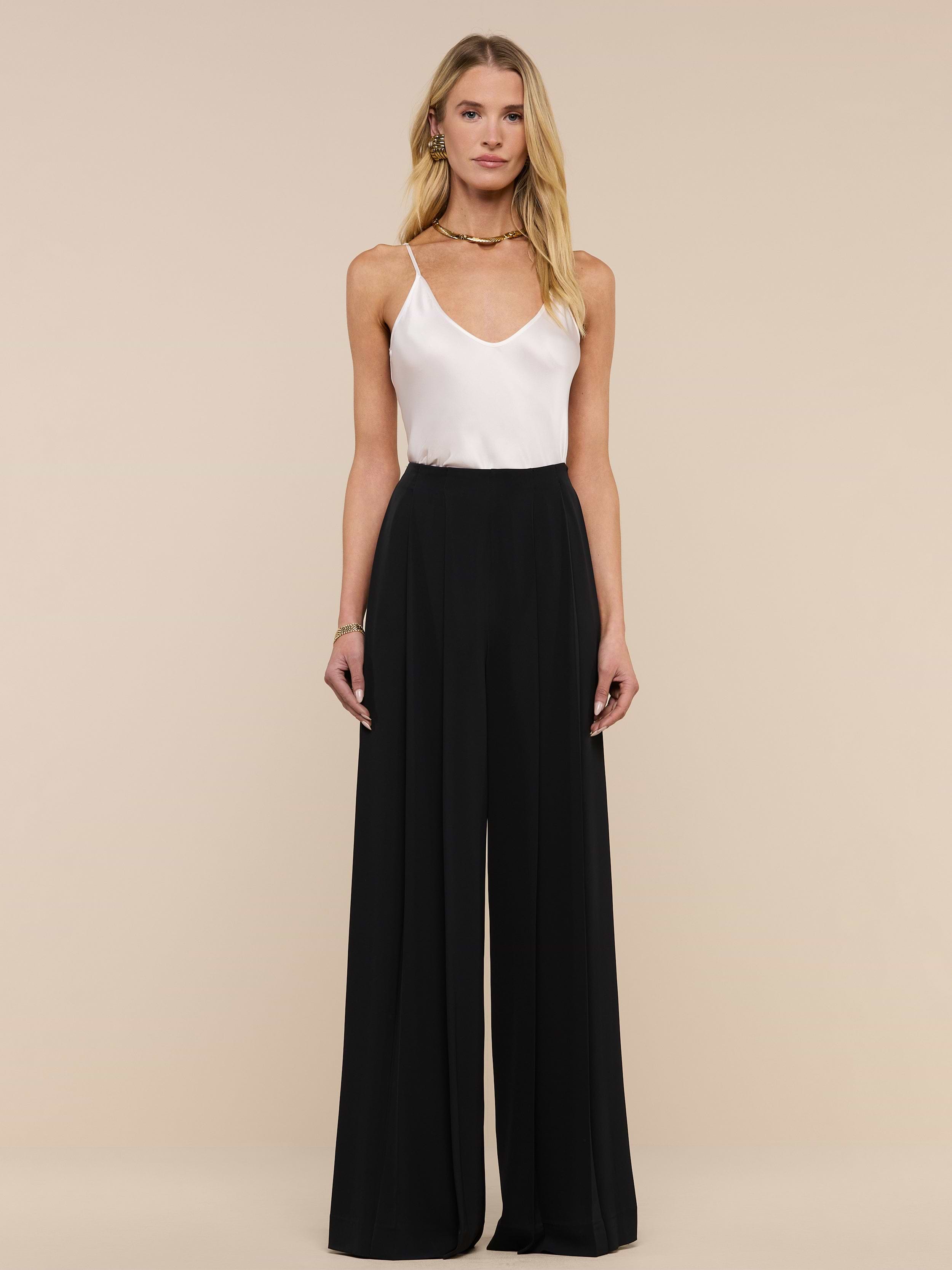 A woman stands against a plain white background, wearing a white sleeveless top paired with the Nova Pleated Wide-Leg Pant, a high-waisted tailored black trouser. Her outfit features wide-leg trousers. She has long blonde hair and a neutral expression.