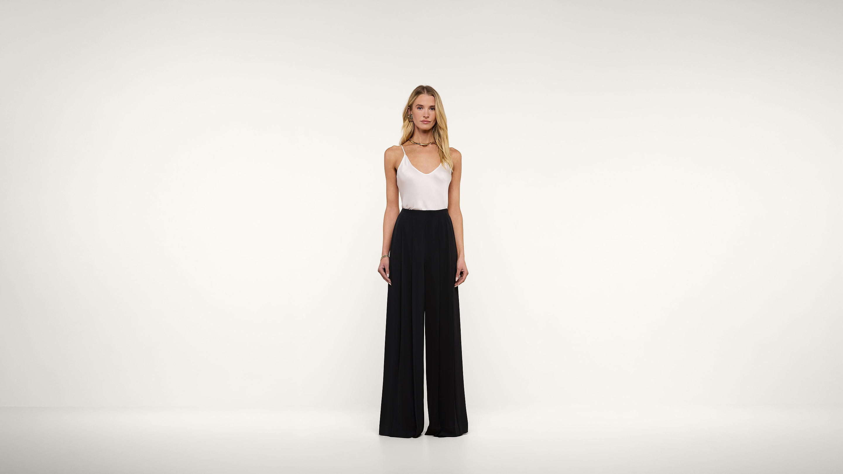 A woman stands in front of a plain white background, wearing a white sleeveless top and high-waisted, wide-leg black pants. She is facing forward with her arms relaxed by her sides.