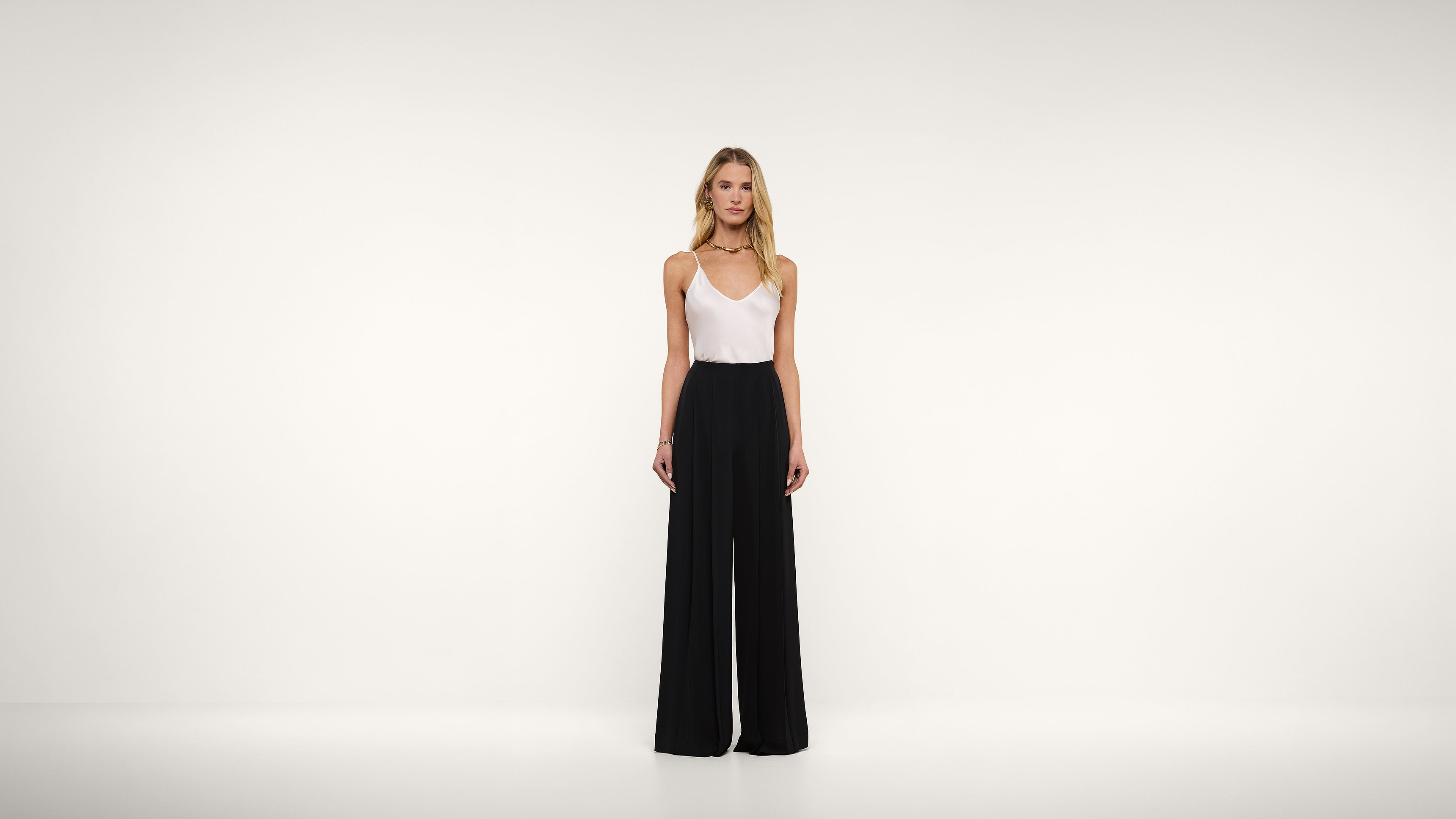 A woman stands in front of a plain white background, wearing a white sleeveless top and high-waisted, wide-leg black pants. She is facing forward with her arms relaxed by her sides.